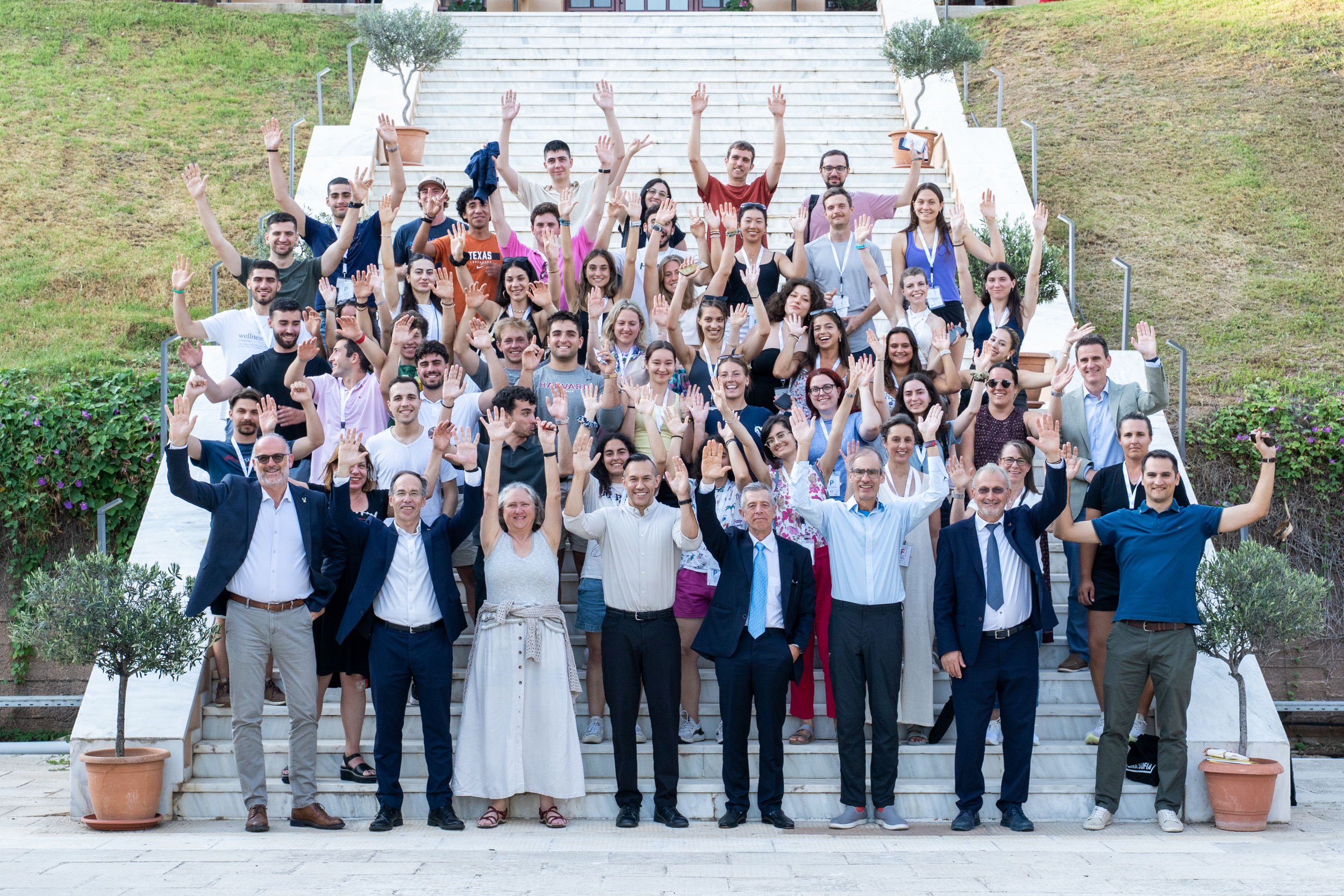 Μια μεγάλη ομάδα ποζάρει σε εξωτερικές σκάλες, χαμογελώντας και σηκώνοντας τα χέρια ψηλά για την ομαδική φωτογραφία.