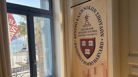 Ένα πανό του Κέντρου Ελληνικών Σπουδών Ελλάδος του Πανεπιστημίου Harvard κρέμεται δίπλα σε ένα παράθυρο με θέα έναν παραθαλάσσιο δρόμο με φοίνικες και σημαίες.