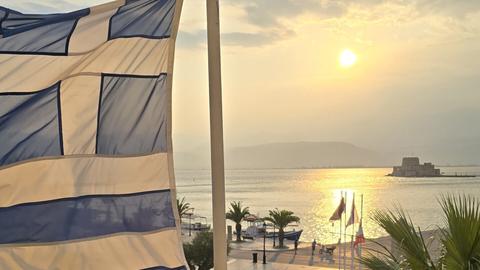 Ελληνική σημαία που κυματίζει πάνω από μια παραλιακή προκυμαία στο ηλιοβασίλεμα, με ανθρώπους να περπατούν από κάτω και τον ήλιο να καθρεφτίζεται στο νερό.