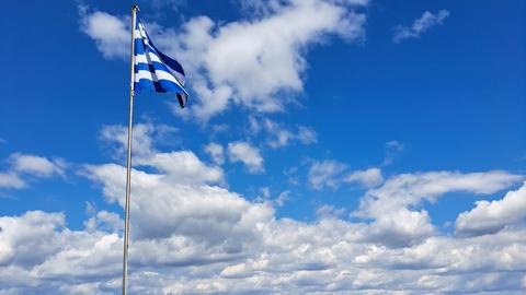 Η ελληνική σημαία κυματίζει σε έναν ιστό πάνω από μια πέτρινη περίφραξη, με θέα τη θάλασσα και τα βουνά κάτω από έναν φωτεινό, μερικώς συννεφιασμένο ουρανό.