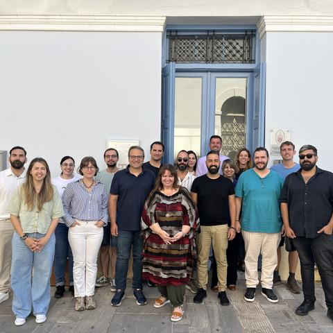 Μια ομάδα ποζάρει για φωτογραφία έξω από το κτήριο του ΚΕΣ με φόντο τα μπλε κουφώματα παραθύρων.