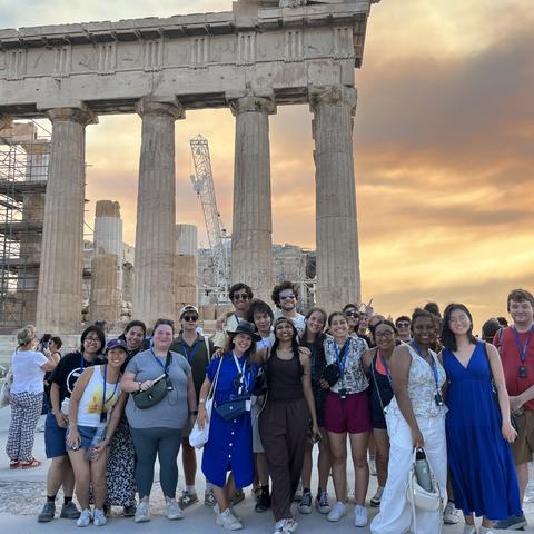 Μια ομάδα ανθρώπων χαμογελά για ομαδική φωτογραφία μπροστά στον Παρθενώνα με φόντο το ηλιοβασίλεμα.