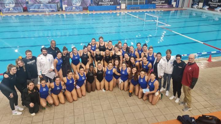 Μια ομάδα νεαρών αθλητριών που φορούν τα χρώματα των ομάδων τους μαζί με τους προπονητές τους ποζάρουν μπροστά από μια εσωτερική πισίνα.