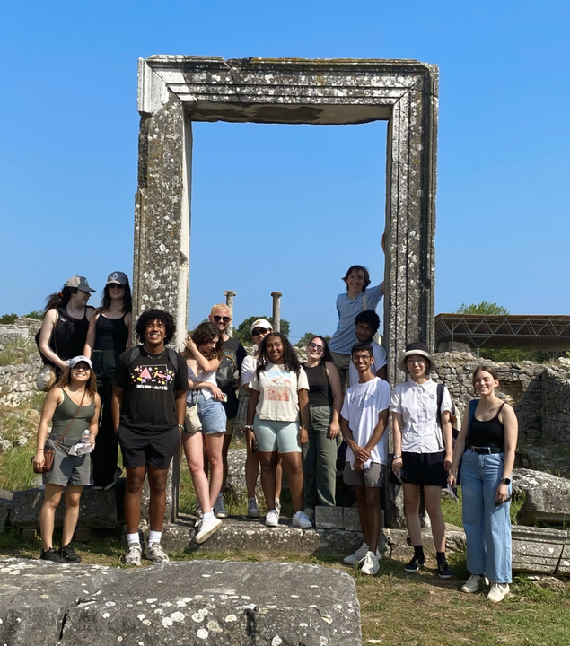 14 νεαρά άτομα ποζάρουν για μια ομαδική φωτογραφία και χαμογελούν στην κάμερα. Στο βάθος διακρίνονται αρχαίες στήλες, ενώ όλοι είναι σκορπισμένοι γύρω από μια αρχαία στήλη σε σχήμα ορθογωνίου.