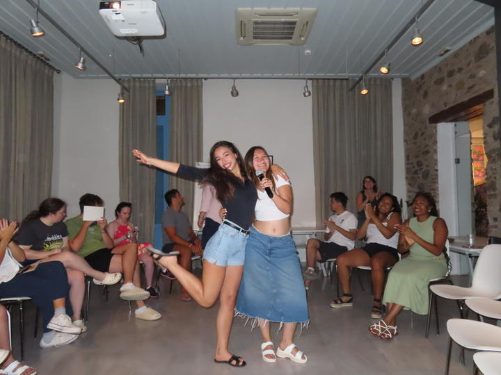 Δύο νεαρές γυναίκες στέκονται και ποζάρουν για την κάμερα στο κέντρο ενός δωματίου με κουρτίνες, πέτρινους τοίχους και φωτισμό οροφής. Η μία κρατά μικρόφωνο, ενώ η άλλη ποζάρει χαρούμενα με το ένα πόδι σηκωμένο. Γύρω τους, μερικά άτομα είναι καθισμένα, χειροκροτούν και χαμογελούν.