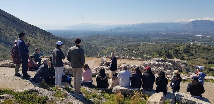 Μια ομάδα είκοσι ατόμων καθισμένων σε κορυφή λόφου ανάμεσα σε αρχαία μνημεία και ακούγοντας έναν άνδρα που στέκεται στη μέση.