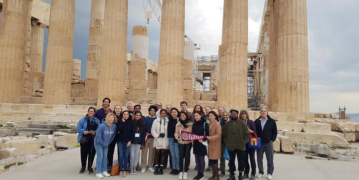 Μια ομάδα είκοσι ατόμων κοιτάζει την κάμερα κρατώντας σημαιάκι του Harvard μπορστά από τον Παρθενώνα στην Αθήνα.