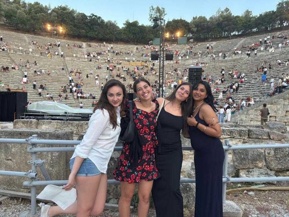Τέσσερις νεαρές γυναίκες ποζάρουν μαζί μπροστά από ένα αρχαίο πέτρινο αμφιθέατρο, χαμογελούν στην κάμερα, ενώ στο βάθος φαίνονται άνθρωποι διασκορπισμένοι στα μεγάλα πετρόκτιστα καθίσματα.