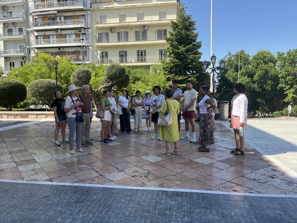 Μια ομάδα ανθρώπων στέκεται σε κύκλο πάνω σε μια πλακόστρωτη πλατεία, περιτριγυρισμένη από δέντρα και πολυκατοικίες, ενώ μια γυναίκα με κίτρινο φόρεμα τούς μιλάει.