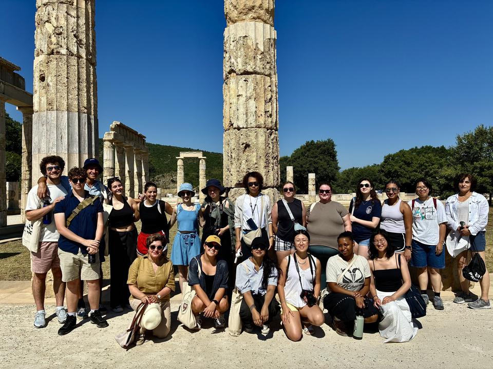 Μια μεγάλη ομάδα ανθρώπων, κάποιοι όρθιοι και κάποιοι καθιστοί, βγάζει αναμνηστική φωτογραφία μπροστά από ψηλές αρχαίες μαρμάρινες κολόνες και ερείπια, κάτω από ένα καθαρό γαλάζιο ουρανό.