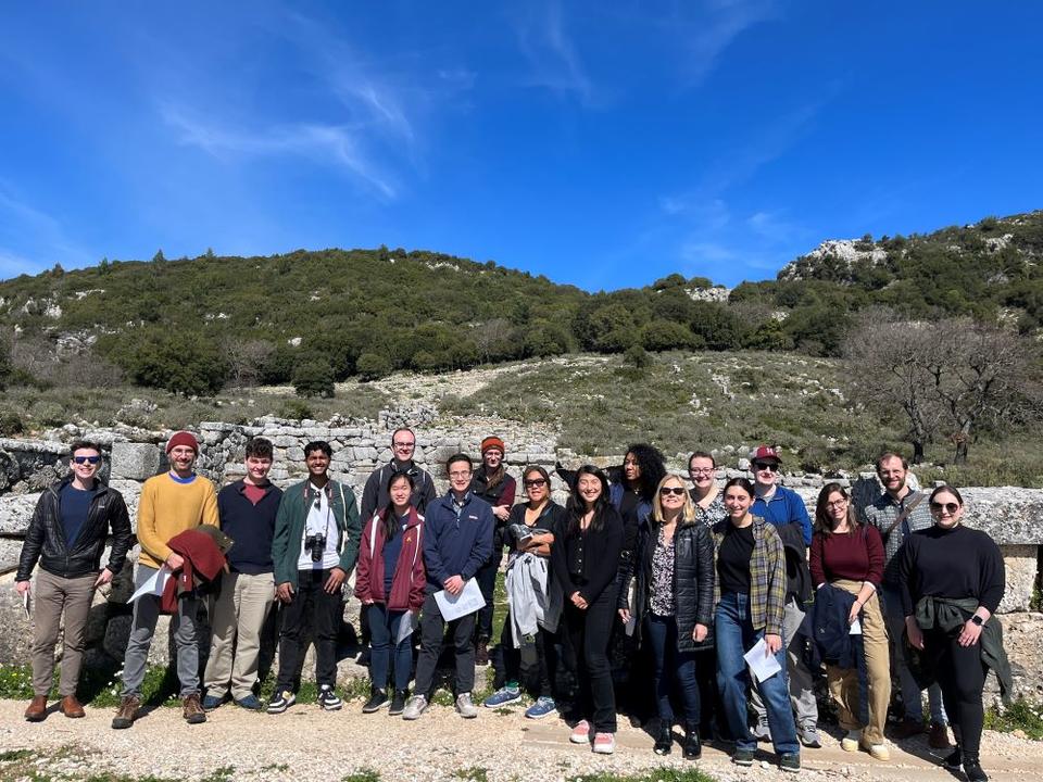 Μια ηλιόλουστη μέρα 18 άτομα στέκονται για ομαδική φωτογραφία μπροστά από αρχαία τείχη και την κορφή μιας πλαγιάς.