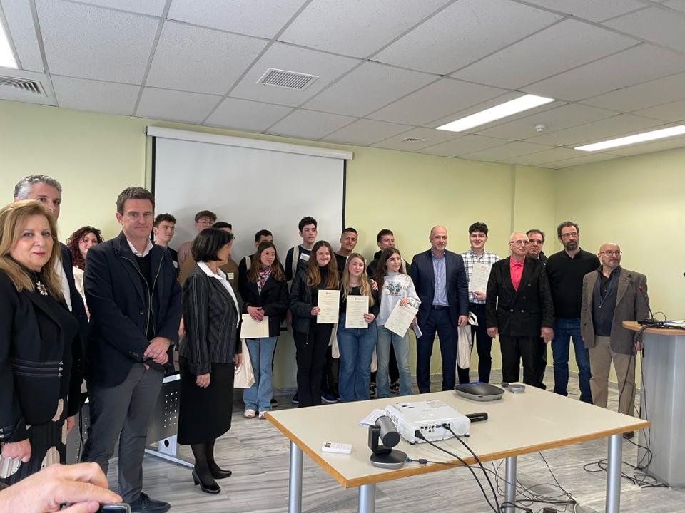 Ομάδα 22 ατόμων διαφόρων ηλικιών χαμογελάει κοιτάζοντας στον φακό σε αίθουσα με πίνακες προβολής στο πίσω μέρος, ενώ ορισμένα νεότερα άτομα κρατάνε βεβαιώσεις της βράβευσής τους.