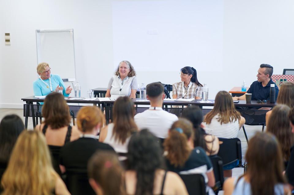 Πάνελ τριών γυναικών και ενός άνδρα σε μια αίθουσα διδασκαλίας. Φαίνεται να συμμετέχουν σε συζήτηση και το κοινό τους ακούει προσεκτικά.