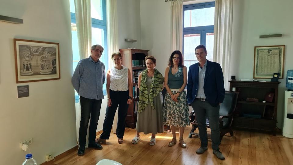 Πέντε άτομα, εκ των οποίων δύο άνδρες στα πλάγια και τρεις γυναίκες στη μέση, στέκονται σε έναν κλειστό χώρο με ξύλινο πάτωμα. Στέκονται ανάμεσα σε παράθυρα απ'όπου μπαίνει φυσικό φως και κοιτούν την κάμερα.