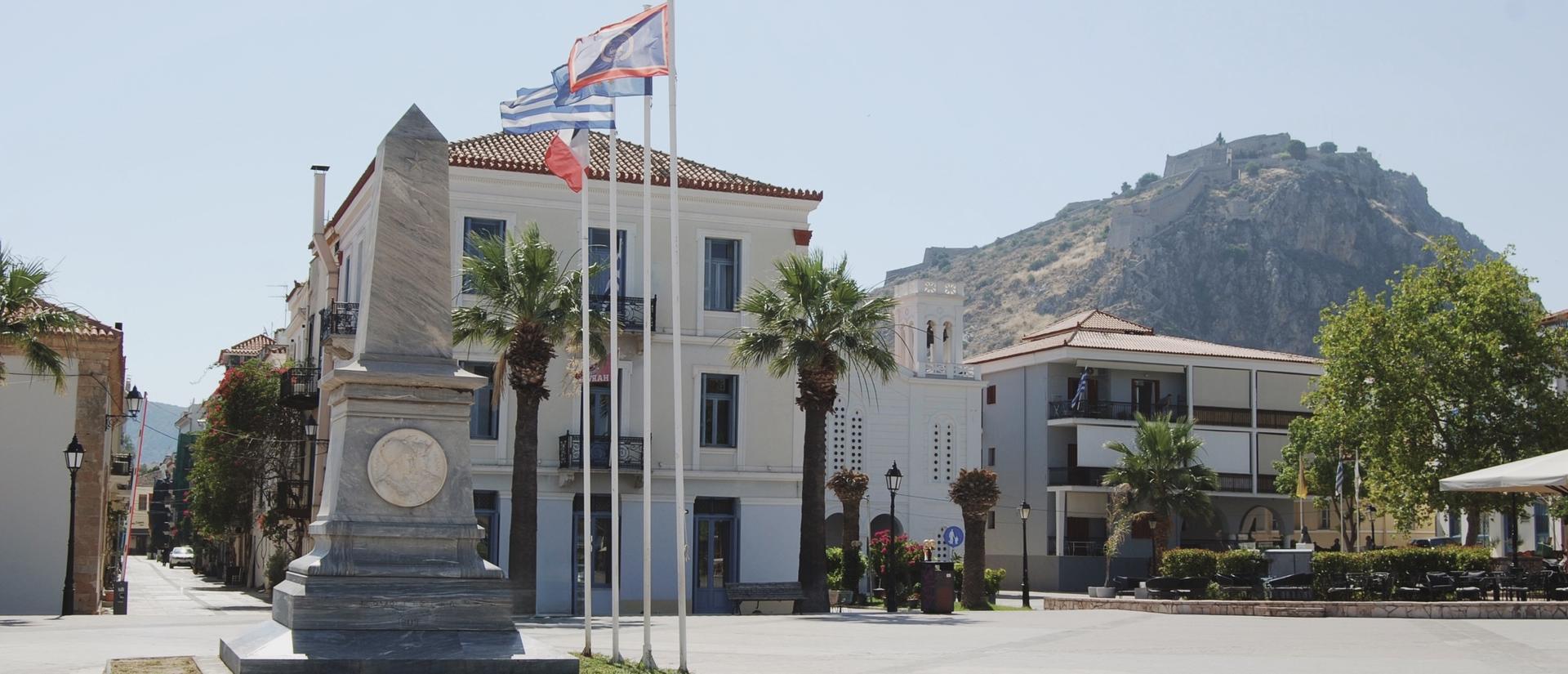 Στο προσκήνιο δεσπόζει ένα μαρμάρινο μνημείο σε μια ηλιόλουστη πλατεία. Πίσω του διακρίνεται το κρεμ κτήριο του ΚΕΣ με μπλε κουφώματα και φοίνικες, ενώ στο βάθος το Παλαμήδι υψώνεται στην κορυφή του λόφου κάτω από καθαρό γαλάζιο ουρανό.