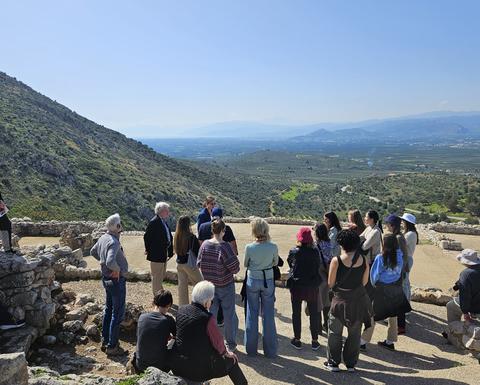 Μια ομάδα στέκεται και κάθεται ανάμεσα σε αρχαία πέτρινα κτίσματα σε μια πλαγιά βουνού, ακούγοντας προσεκτικά μια γυναίκα που μιλά.