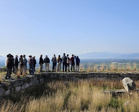 Μια ομάδα ανθρώπων έχει συγκεντρωθεί πάνω σε απομεινάρια μιας αρχαίας πέτρινης κατασκευής, αγναντεύοντας μια εκτεταμένη κοιλάδα με διάσπαρτους οικισμούς και μακρινά βουνά κάτω από έναν καταγάλανο ουρανό.