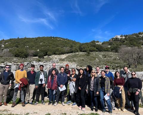 Μια ηλιόλουστη μέρα 18 άτομα στέκονται για ομαδική φωτογραφία μπροστά από αρχαία τείχη και την κορφή μιας πλαγιάς.