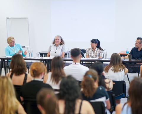 Πάνελ τριών γυναικών και ενός άνδρα σε μια αίθουσα διδασκαλίας. Φαίνεται να συμμετέχουν σε συζήτηση και το κοινό τους ακούει προσεκτικά.